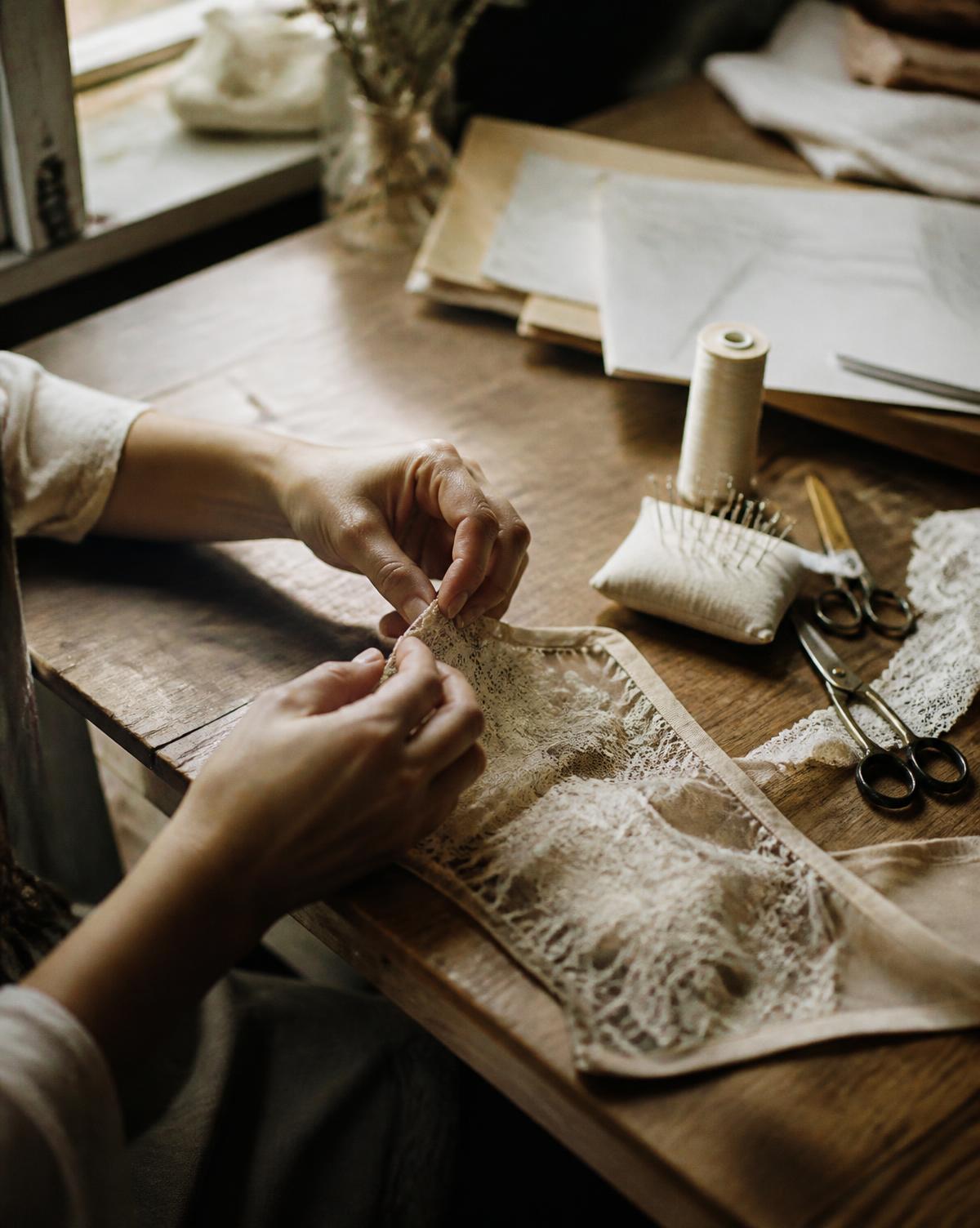 Hands stitching lace at a wooden worktable in natural light